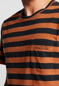Acercamiento a una camisa a rayas marrón y negra con un bolsillo en el pecho etiquetado "AMS Couture Scotch & Soda KG 22."