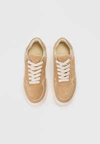 Baskets en suede beige avec des lacets crème, un col rembourré et une semelle en caoutchouc blanche. Présente des accents de couture et une languette de marque.