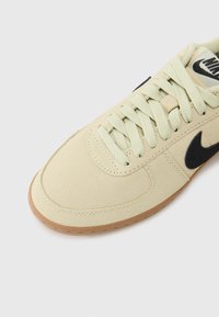 Lys beige sneaker med en suede-textur; har flade grønne snørebånd, et sort logo og en sål af gummirubber. Buet tådesign.