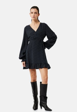 Nainen pukeutunut mustaan pitkähihainen V-kaula-aukkoiseen minimekkoon, jossa on pieniä valkoisia pilkkuja, röyhelöhelma ja mustat polven korkeudelle ulottuvat korolliset saappaat.