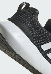 Scarpa da ginnastica nera e grigia in maglia con suola in gomma bianca e tre strisce bianche. Presenta una parte superiore testurizzata e un tassello di presa sul tallone.