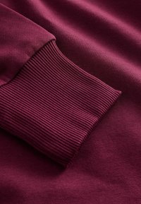 Burgundská svetr látka s žebrovaným manžetou. Hladká textura na těle, kontrastní žebrování a jednoduchý design.