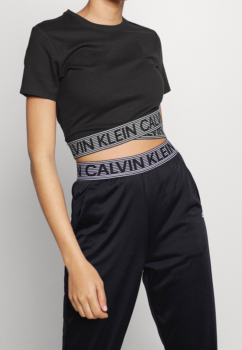Personne portant un t-shirt court noir et un pantalon noir avec des bandes élastiques visibles à la marque Calvin Klein sur les deux vêtements.