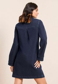Femme aux cheveux sombres mi-longs portant une robe en denim bleu marine à manches longues, se tenant avec une main sur la hanche, tournant le dos à l'appareil photo.
