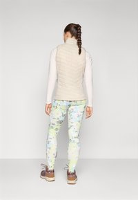 Lys beige quiltet vest over en langærmet hvid top, parret med pastelfarvede camouflagemønstrede leggings og brune vandresko. Fletningen i håret er synlig.