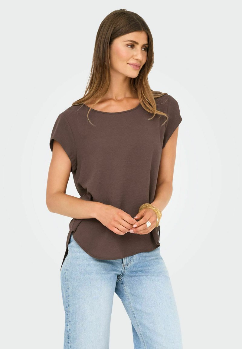 Donna con lunghi capelli castani che indossa una maglietta marrone a maniche corte e jeans azzurri chiari, in piedi con le mani leggermente intrecciate, che guarda verso destra.
