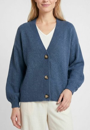 Femme portant un cardigan en tricot bleu à trois boutons par-dessus un haut blanc et un pantalon clair, debout devant un fond uni.