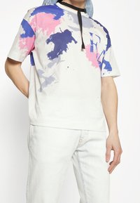 Krátký rukáv, bílý tričko s barevnými abstraktními skvrnami barvy růžové, modré a šedé; má detail černé kravaty. Viditelné džínové kalhoty.