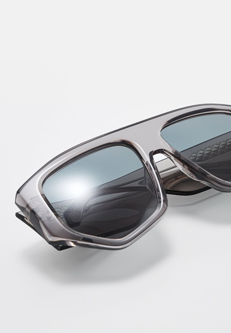 Lunettes de soleil rectangulaires surdimensionnées et translucides grises avec des verres teintés foncés reposant sur une surface blanche.