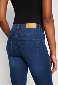 Jeans de ganga azul com um corte slim, apresentando uma etiqueta de pele na cintura, detalhes de costura subtis e bolsos traseiros.