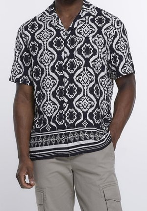 Homme portant une chemise à manches courtes boutonnée à motifs noirs et blancs avec un short cargo beige, une main dans la poche.