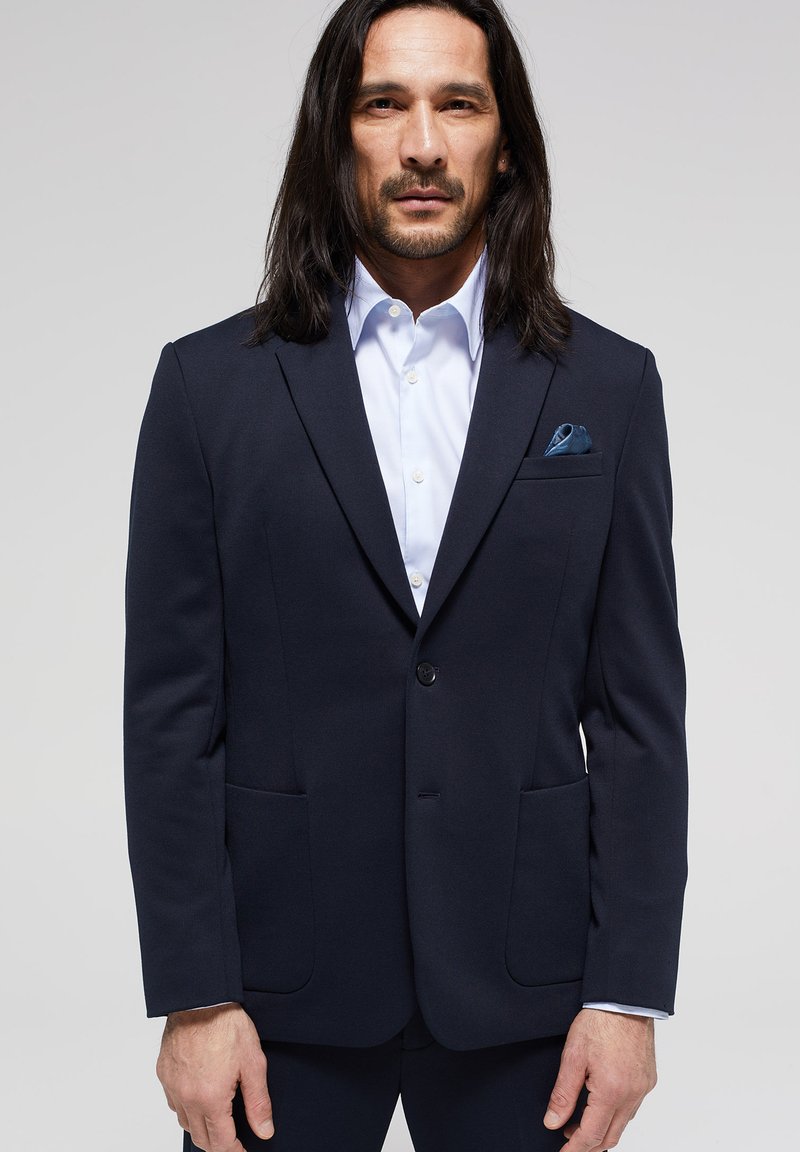 Hombre con cabello largo y oscuro vestido con una chaqueta de traje azul marino, camisa de vestir blanca y un pañuelo azul en el bolsillo, de pie frente a un fondo sencillo.