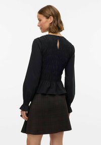 Sort bluse med smockdetalje på ryggen, lange ærmer og peplumkant, kombineret med et ternet nederdel i mørke nuancer. Bagfra set med nøglehulsåbning.