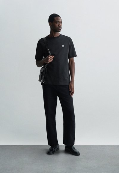 Tricou negru cu mânecă scurtă, având un mic emblemă grafică, asortat cu pantaloni largi negri și pantofi negri, purtând o geantă tip crossbody.