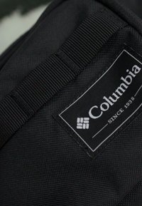 Sort nylon rygsæk hjørne med et mærke, der har "Columbia" og "SINCE 1938" tekst, omkranset af tekstureret stof og webbing-detaljer.