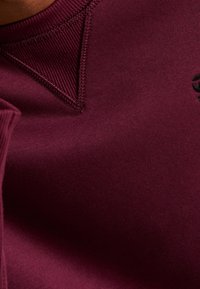 Primo piano di un tessuto bordeaux con scollo a costine e un elemento di design triangolare. Texture liscia con una leggera lucentezza e senza motivi visibili.