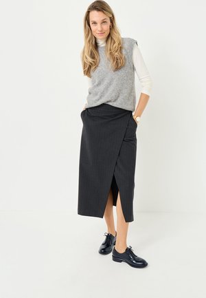 Maglione grigio senza maniche sopra una maglietta bianca a maniche lunghe, abbinato a una gonna avvolgente nera a righe sottili con uno spacco laterale e scarpe nere.
