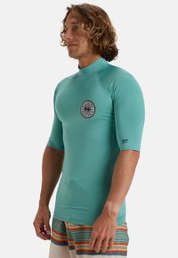 Rash guard de manga corta en color teal con un logo redondo en el pecho. Hecho de un material suave y elástico, diseñado para deportes acuáticos.