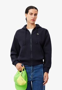 Navy zip-up hoodie with drawstrings and kangaroo pockets, pair with light blue denim jeans, holding a green cap with perforations.