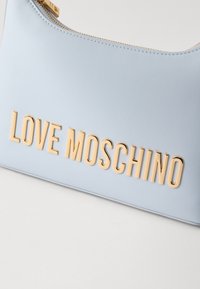 Svetlo modra torba iz sintetičnega usnja z zlatimi vtisnjenimi črkami "LOVE MOSCHINO", gladka tekstura in zadrgo na vrhu.