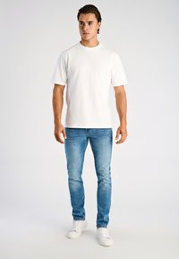 Weißes Baumwoll-T-Shirt mit kurzen Ärmeln, kombiniert mit hellblauen Jeans. Das Model trägt weiße Sneakers und steht vor einem schlichten weißen Hintergrund.