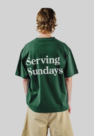 Person mit welligem Haar, die ein dunkelgrünes T-Shirt mit weißem Aufdruck "Serving Sundays" trägt, von hinten vor einem grauen Hintergrund zu sehen.