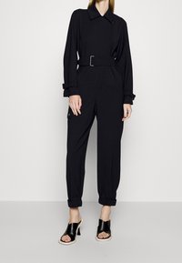 Mono negro con cinturón, de manga larga, ajuste entallado, bolsillos laterales, dobladillos con puños, y combinado con tacones negros con tiras. Textura de tela suave.