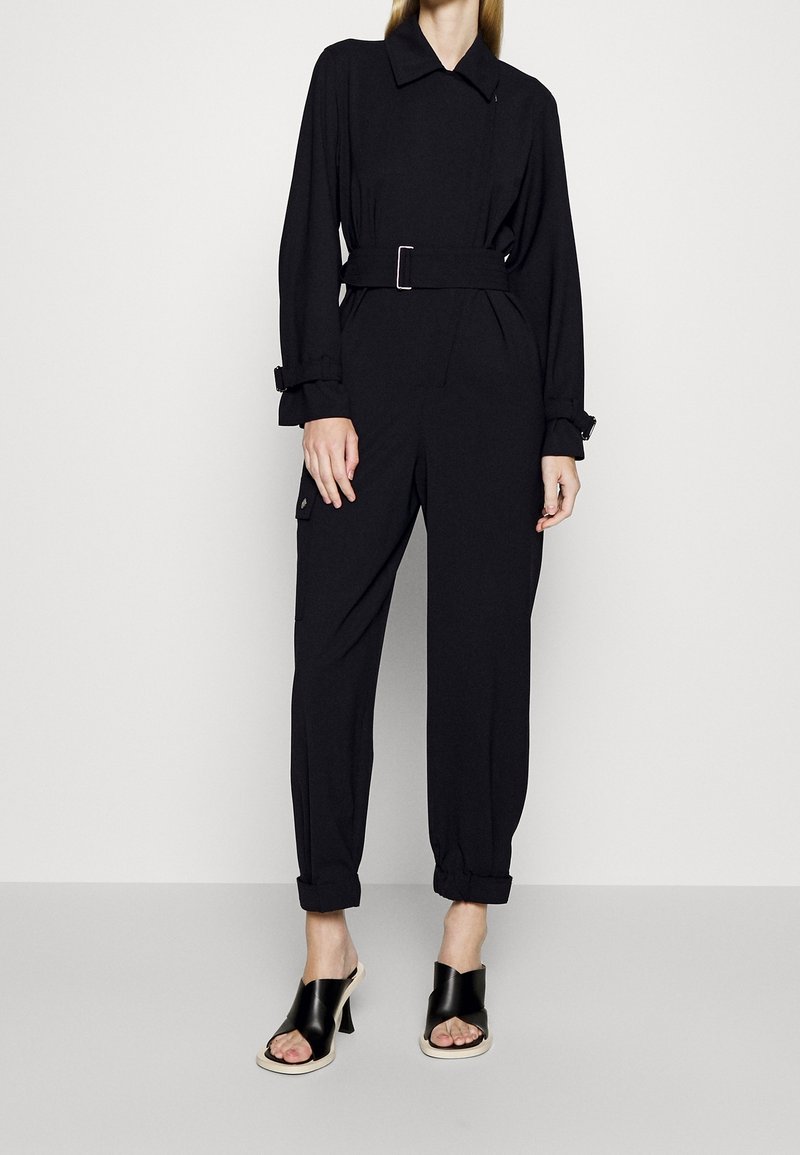 Mono negro con cinturón, de manga larga, ajuste entallado, bolsillos laterales, dobladillos con puños, y combinado con tacones negros con tiras. Textura de tela suave.