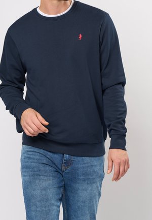 Sudadera de algodón azul marino con cuello redondo de canalé, mangas largas y pequeño logo rojo en el pecho. Combinada con jeans de mezclilla azul claro.
