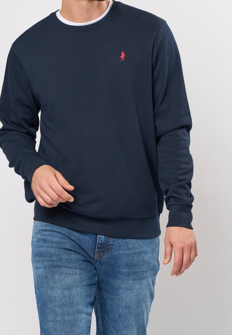 Felpa in cotone blu navy con collo a girocollo a costine, maniche lunghe e piccolo logo rosso sul petto. Abbinata a jeans in denim azzurro.