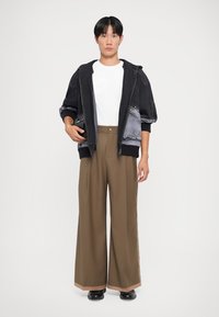 Black denim oversized jacket with a gray gradient, paired with wide brown trousers and black shoes. White t-shirt underneath, hands on pockets.