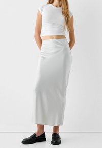 Bílý crop top s krátkými rukávy, zkombinovaný s dlouhou, volnou, bílou saténovou sukní. Černé mokasíny s texturovanými detaily dotvářejí outfit.