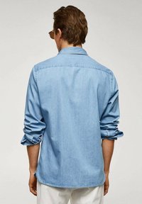 Chemise en denim bleu clair à manches longues, col boutonné et poignets retroussés, avec des détails de couture minimalistes et une coupe droite.