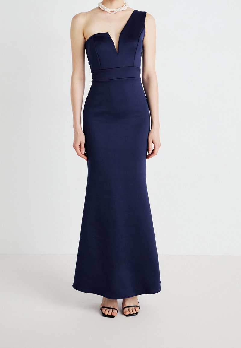 Femme portant une robe de soirée bleu marine asymétrique avec un décolleté en V profond, associée à un collier blanc perlé et des talons noirs à brides.