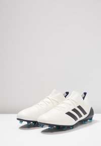 Botas de fútbol blancas con tres rayas negras, un material superior liso y taches azules translúcidos sobre una suela negra. Diseño de corte bajo.