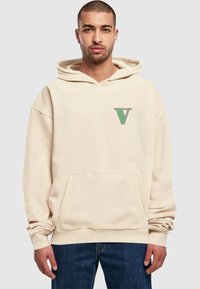 Béžová mikina s veľkou prednou kapsou, zeleným logom "V" na hrudi a voľným strihom. Vyrobená z mäkkej tkaniny s dlhými rukávmi.