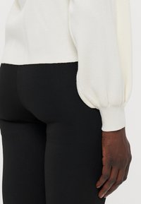 Pull blanc avec des manches bouffantes et des poignets côtelés, associé à un pantalon noir ajusté. La texture est lisse et le tissu semble doux.