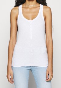 Vit linne med rundad halsringning, knappstängning framtill, tillverkad av lätt tyg med en lätt textur. Avslappnad passform, ärmlös design.