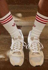 Ben som bär vita Adidas-strumpor med röda ränder och beige Adidas-sneakers med vita snörning, placerade på betongtrappor.