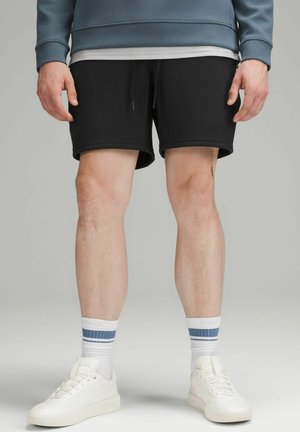 Sorte atletikshorts med snøre, kombineret med hvide sneakers og stribede sokker. Shortsene har en glat tekstur og midt på låret længde.