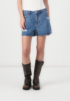 Person iført blå cowboyshorts med let slid og sorte knælange støvler, stående mod en ensfarvet hvid baggrund.
