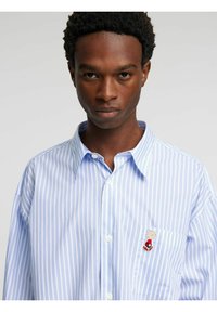 Srajca z gumbi z navpičnimi svetlo modrimi in belimi črtami ter majhno izvezeno grafiko na prsnem žepu. Bombažni material.