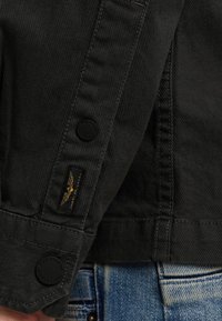 Manche de veste en denim noir avec des détails de couture, ornée d'un bouton noir et d'une étiquette avec un logo d'aigle. Portée sur un jean bleu déchiré.