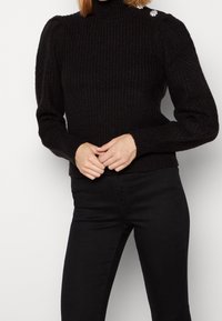 Maglione a costine nero con collo alto, dotato di maniche a sbuffo e un accento floreale decorativo sulla spalla. Indossato con pantaloni neri.