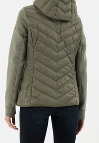 Giacca ibrida verde oliva con cappuccio trapuntato, cuciture a zigzag sul retro e materiale morbido elasticizzato sulle maniche e sui lati.