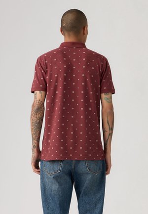 Homme tatoué portant un polo bordeaux à manches courtes avec de petits motifs carrés blancs et un jean en denim bleu, de dos.