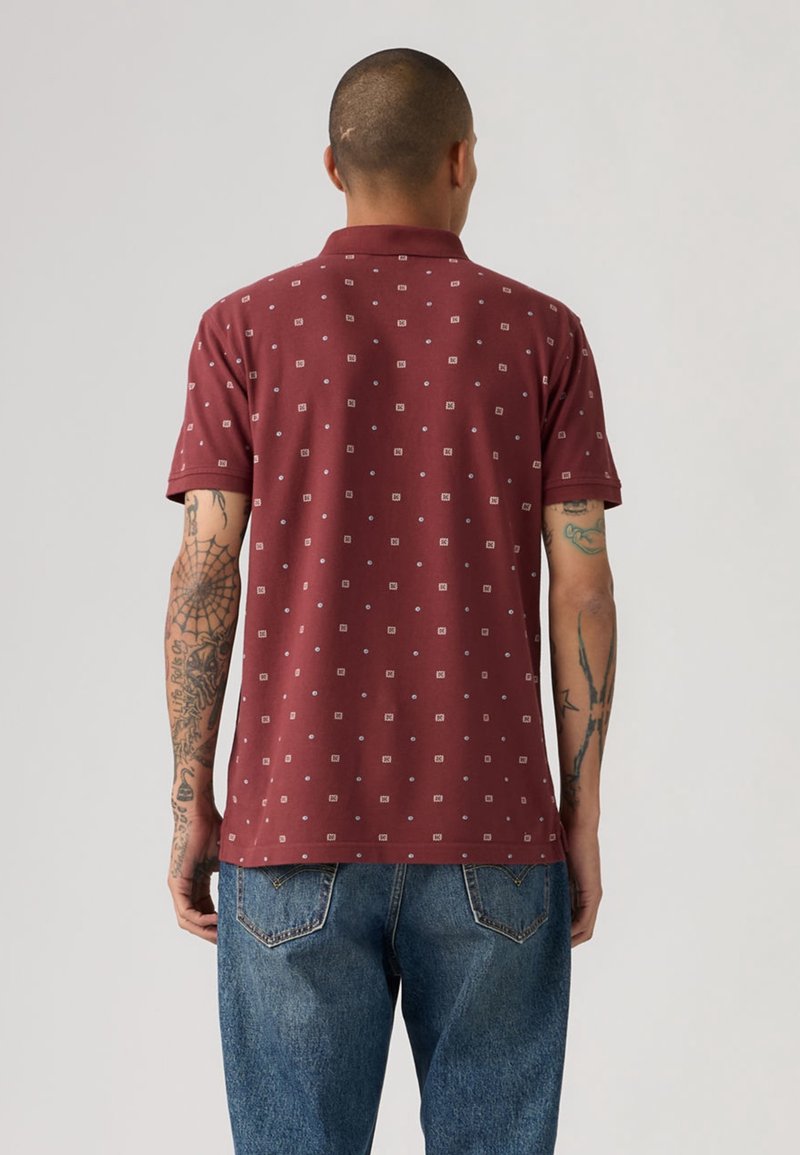 Homme tatoué portant un polo bordeaux à manches courtes avec de petits motifs carrés blancs et un jean en denim bleu, de dos.
