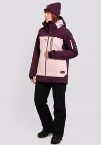 Purple and pink outdoor jacket with zippered pockets, hood, and adjustable cuffs, paired with black pants and dark footwear.