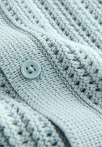 Pull en tricot bleu clair avec un motif texturé. Présente un bouton rond bleu et une apparence douce et duveteuse.