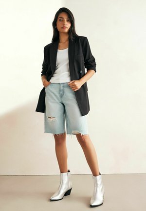 Blazer negro, camiseta sin mangas blanca, bermudas desgastadas de color azul claro y botas de tobillo plateadas metálicas con un acabado texturizado.
